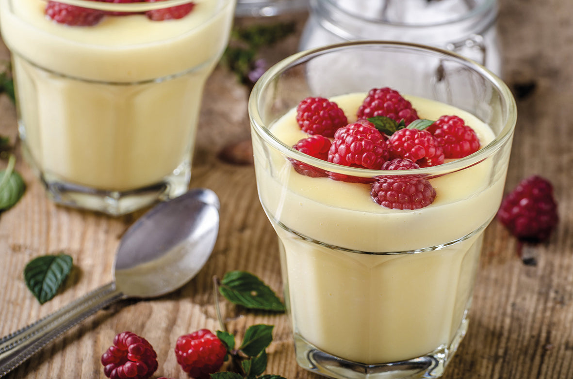 Zwei Gläser mit cremigem Vanillepudding, dekoriert mit frischen Himbeeren und Minzblättern auf rustikalem Holz