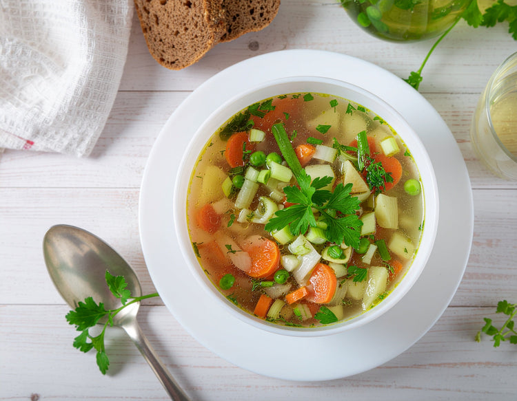 Teller mit klarer Gemüsesuppe, gefüllt mit Karotten, Erbsen, Sellerie und Kartoffelstücken, garniert mit frischer Petersilie, daneben ein Löffel und eine Scheibe Brot auf hellem Holztisch. Beispielbild für unsere Kategorie "Brühen & Suppen pflanzlich".