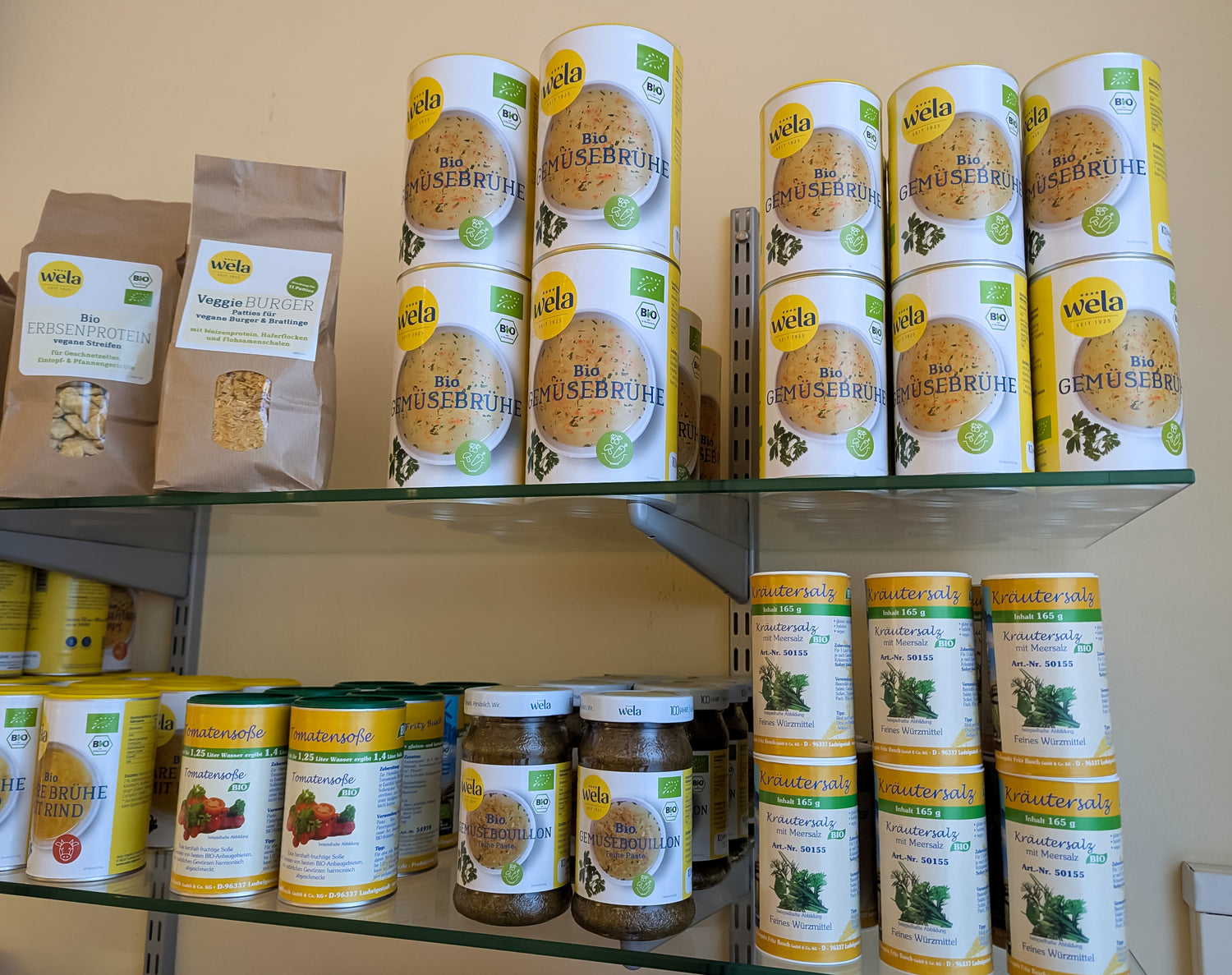 Regal mit Wela Bio-Gemüsebrühe, Kräutersalz und Bio Produkten in Dosen und Beuteln in einer Verkaufsumgebung.